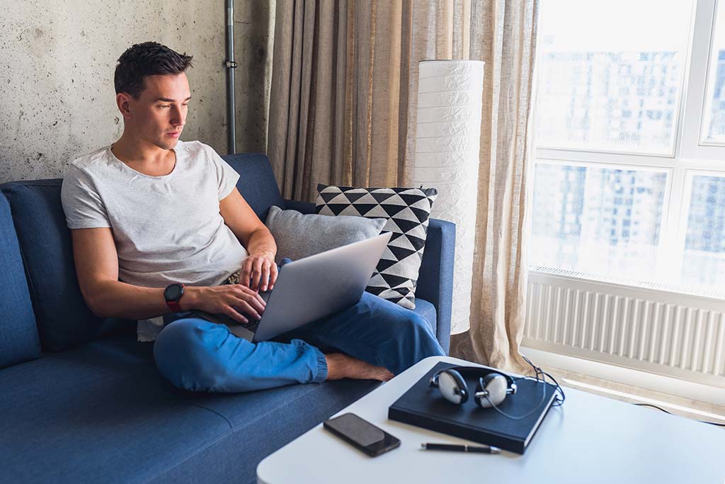 یک پسر جوان که با لباس راحتی روی مبل نشسته و در حال کار رکدن با لپ تاپ است