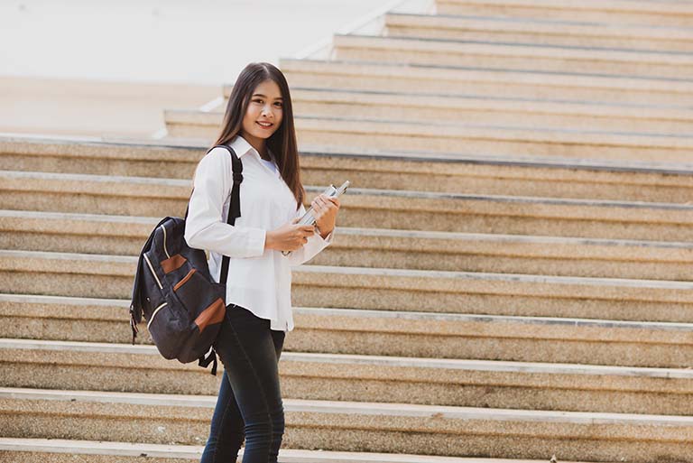 دختر دانشجو در حالی که کوله لپ تاپ را بر دوش خود دارد و در کنار پله ها ایستاده، وزن بالای کوله به خوبی حس می‌شود